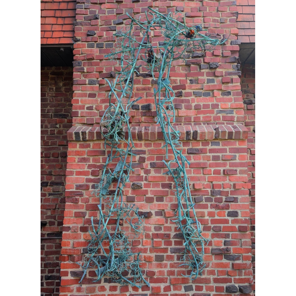 A large, abstract turquoise metal sculpture resembling tangled vines or branches is mounted vertically against a red brick wall. The elongated form features twisting rods, curled wire-like details, and scattered leaf-like elements.