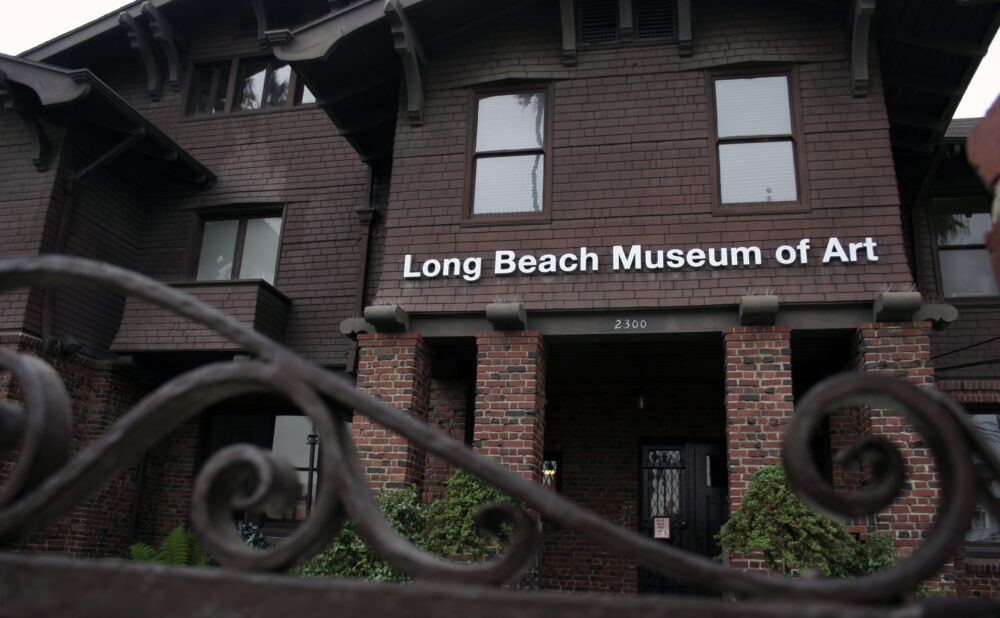 Exterior view of the Long Beach Museum of Art building, a dark brown brick structure with a sign reading “Long Beach Museum of Art” above the entrance. A decorative wrought-iron fence is visible in the foreground, with brick columns, shrubs, and the museum entrance in the background under overcast daylight.