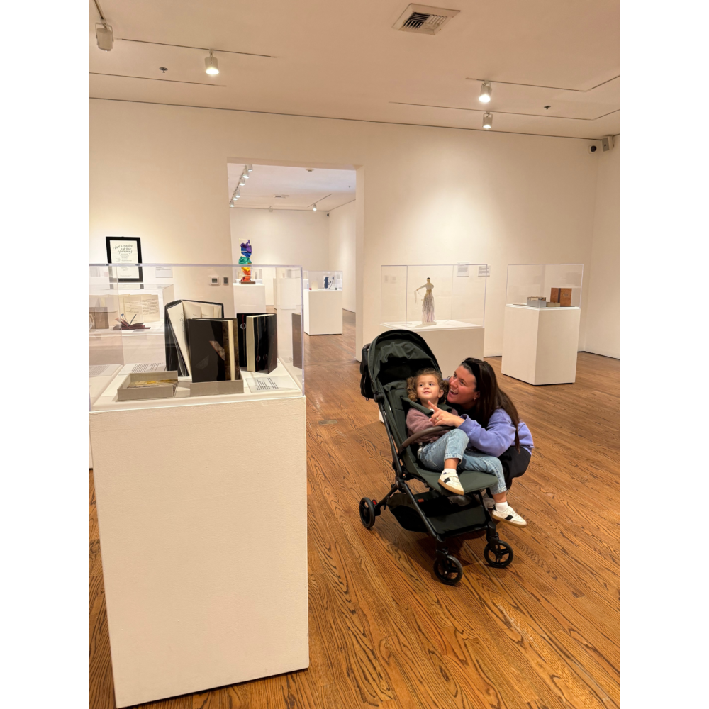 An adult kneels beside a child in a stroller, gently holding and talking with them inside a quiet art gallery filled with display pedestals.