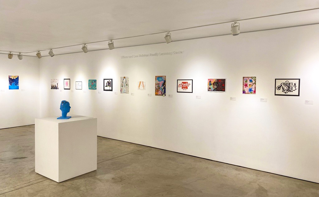 A gallery view with a row of colorful framed artworks displayed along a white wall, and a bright blue sculpted head on a pedestal in the foreground.