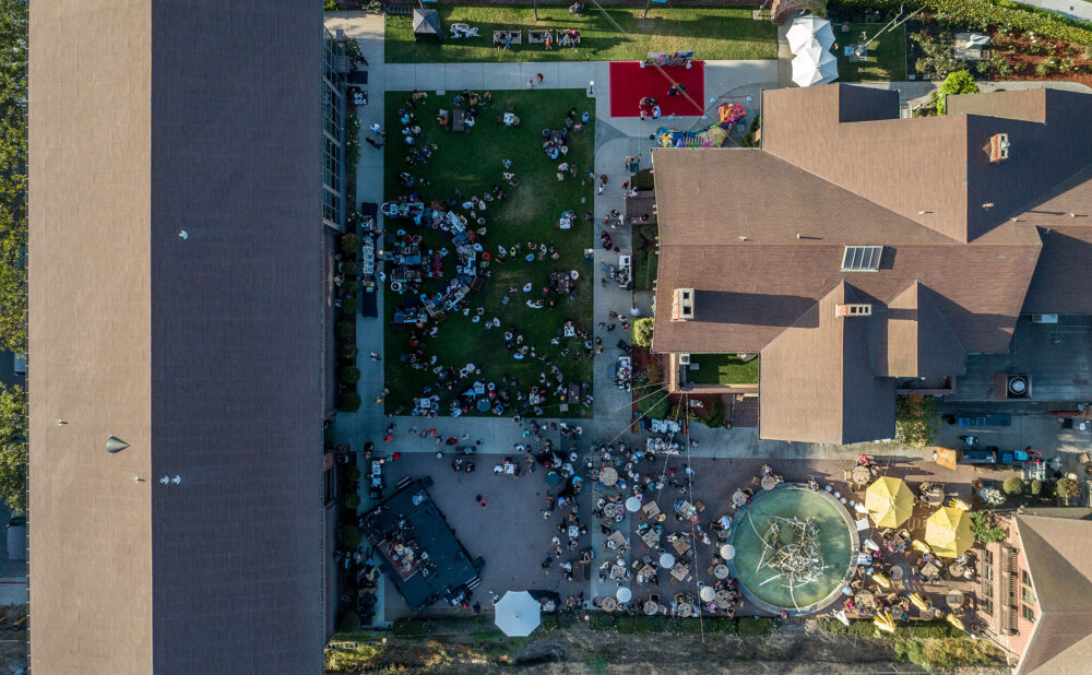 Aerial view of an LBMA's courtyard event with groups of people seated at tables, green lawn areas, a red carpet walkway, and a central fountain.