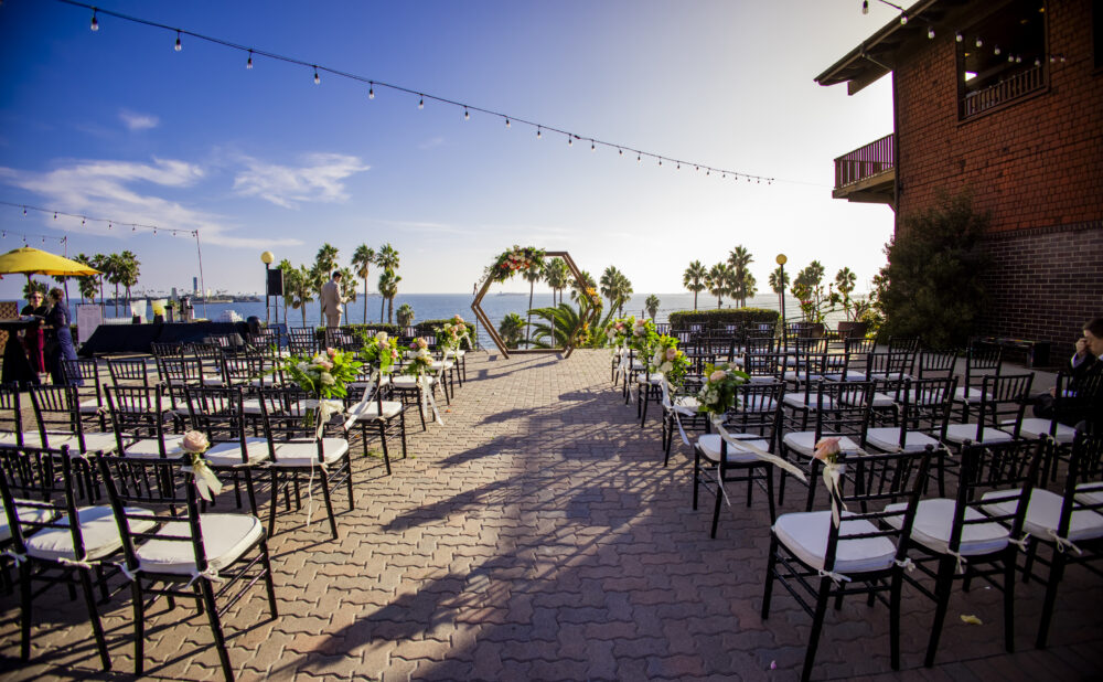 Outdoor event setup with rows of black chairs arranged along a brick walkway, floral arrangements lining the aisle, and a decorative arch at the front. Palm trees and the ocean are visible in the background under string lights at sunset.
