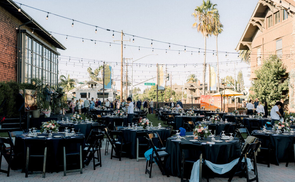 Outdoor event setup with round banquet tables arranged under string lights as guests arrive and walk through the space.