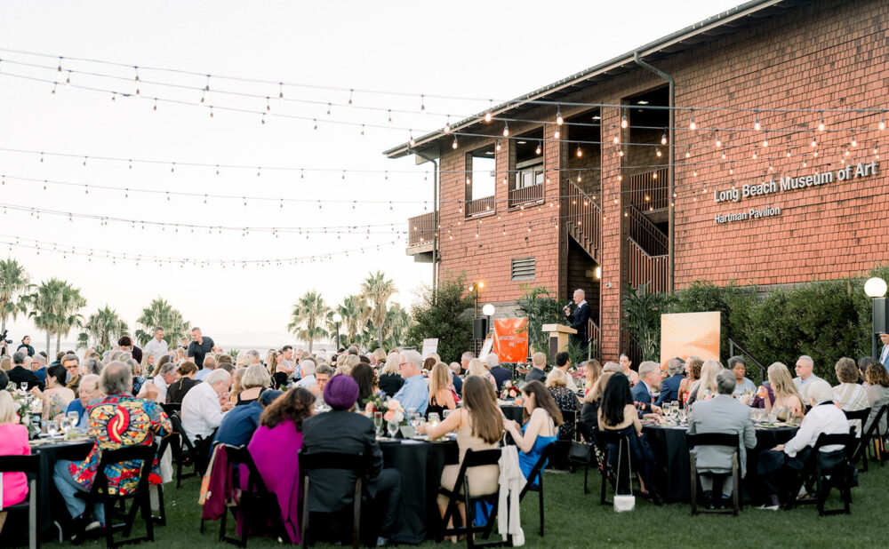 Outdoor evening event with round banquet tables and seated guests on a lawn in front of a brick building labeled ‘Long Beach Museum of Art.’ String lights hang overhead, with palm trees visible in the background.