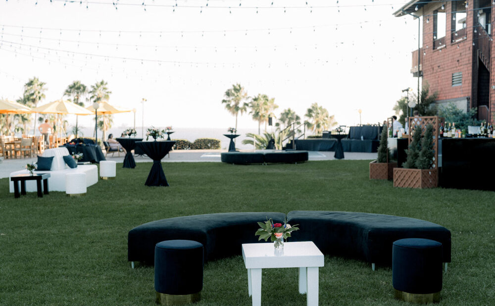 Outdoor lounge area set on a grassy lawn with black and white seating, cocktail tables, and string lights overhead. Palm trees and the ocean are visible beyond the lawn, beside a brick building.