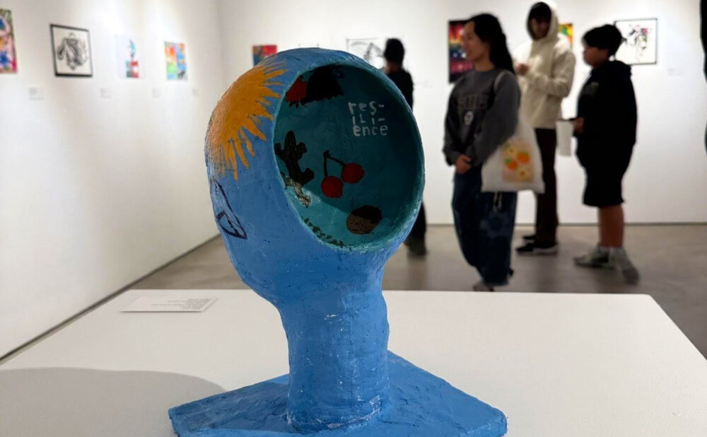 A painted blue head sculpture with an open, bowl-like top displaying illustrations and the word “resilience,” shown on a pedestal in a gallery with visitors viewing artwork in the background.