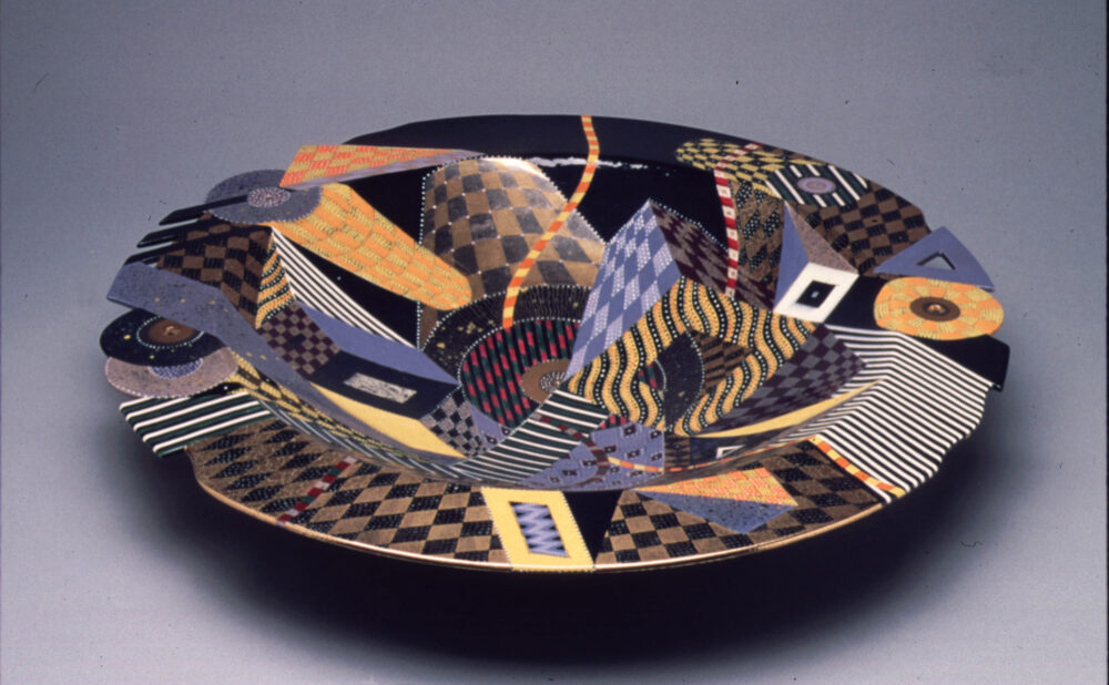 A wide, shallow ceramic bowl with a black interior and an intricate arrangement of overlapping geometric patterns, including checkerboards, stripes, diamonds, and circular motifs. The design features black, gold, orange, lavender, red, and white, creating a layered, abstract composition against a gray background.