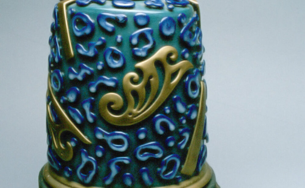 A cylindrical ceramic sculpture with a turquoise-green body covered in raised cobalt-blue abstract shapes and gold decorative flourishes. A small figurative ornament and two gold circular forms crown the blue lid. The sculpture rests on a dark green, textured base against a pale blue background.