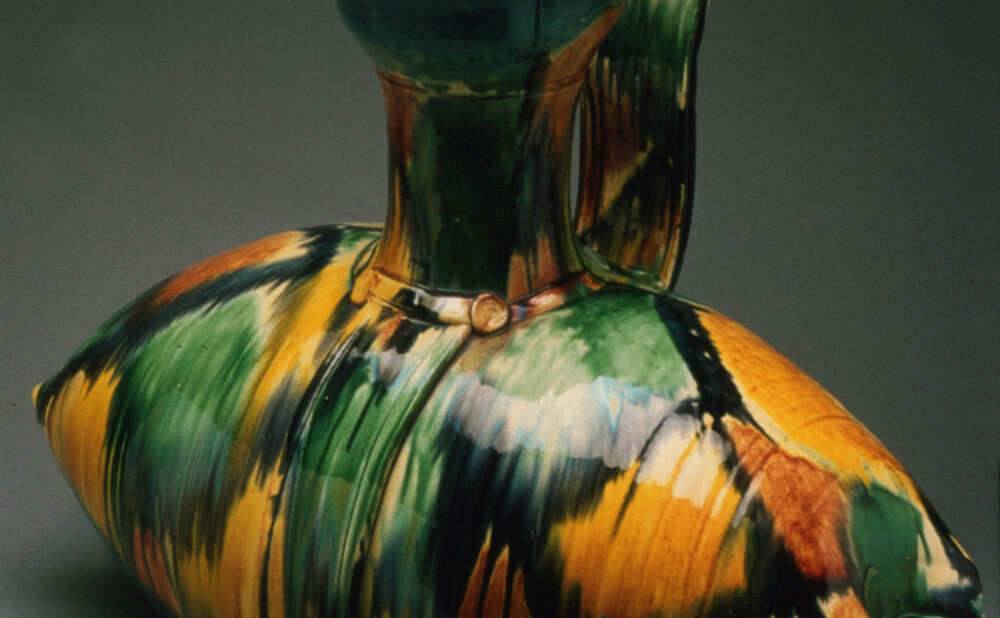 A sculptural ceramic vessel with a wide, rounded body, curved spout, and tall, looped handle. Its glossy surface features bold, expressive brushstrokes of turquoise, green, yellow, orange, black, and white against a dark gray background.