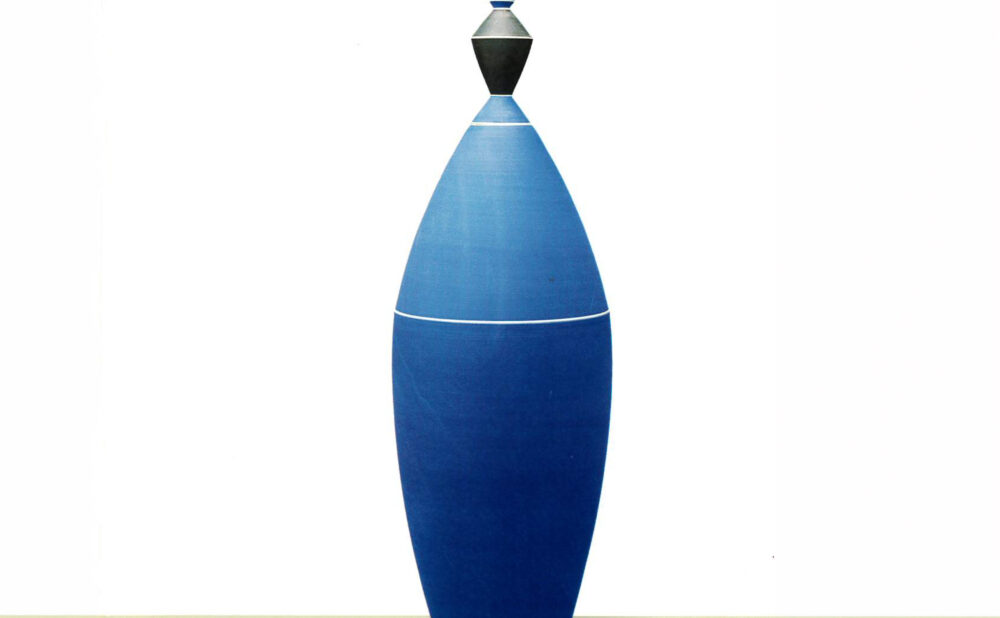A tall, slender ceramic sculpture with an elongated, tapered blue body, accented by thin white horizontal lines. The sculpture is topped with a small black-and-silver geometric form and a pointed blue finial, standing against a white background.
