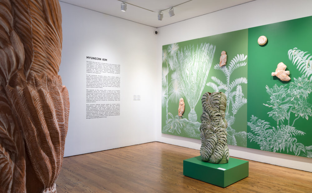 A museum gallery with wood floors and white walls featuring a large green mural of stylized plants and foliage. In front of the mural, a textured, organic-shaped sculpture sits on a green pedestal, while another tall, carved wooden sculpture stands in the foreground. Wall text introducing the artist Myungjin Kim is visible to the left.