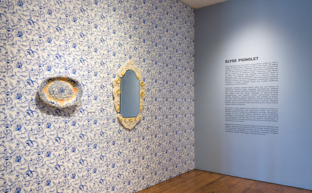 A gallery corner with floor-to-ceiling blue-and-white patterned wallpaper. Two decorative ceramic works—a shallow platter and an ornate framed mirror—are mounted on the wall. Adjacent wall text introduces the artist Elyse Pignolet.