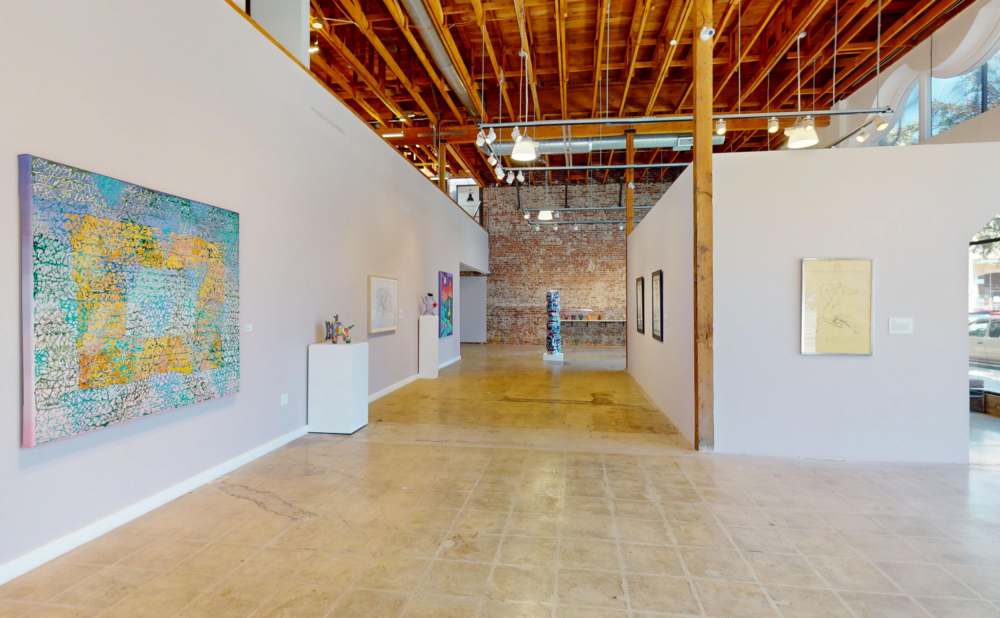 Interior view of the Long Beach Museum of Art Downtown galleries, featuring contemporary artworks displayed on white walls beneath an exposed wood-beam ceiling and brick wall.