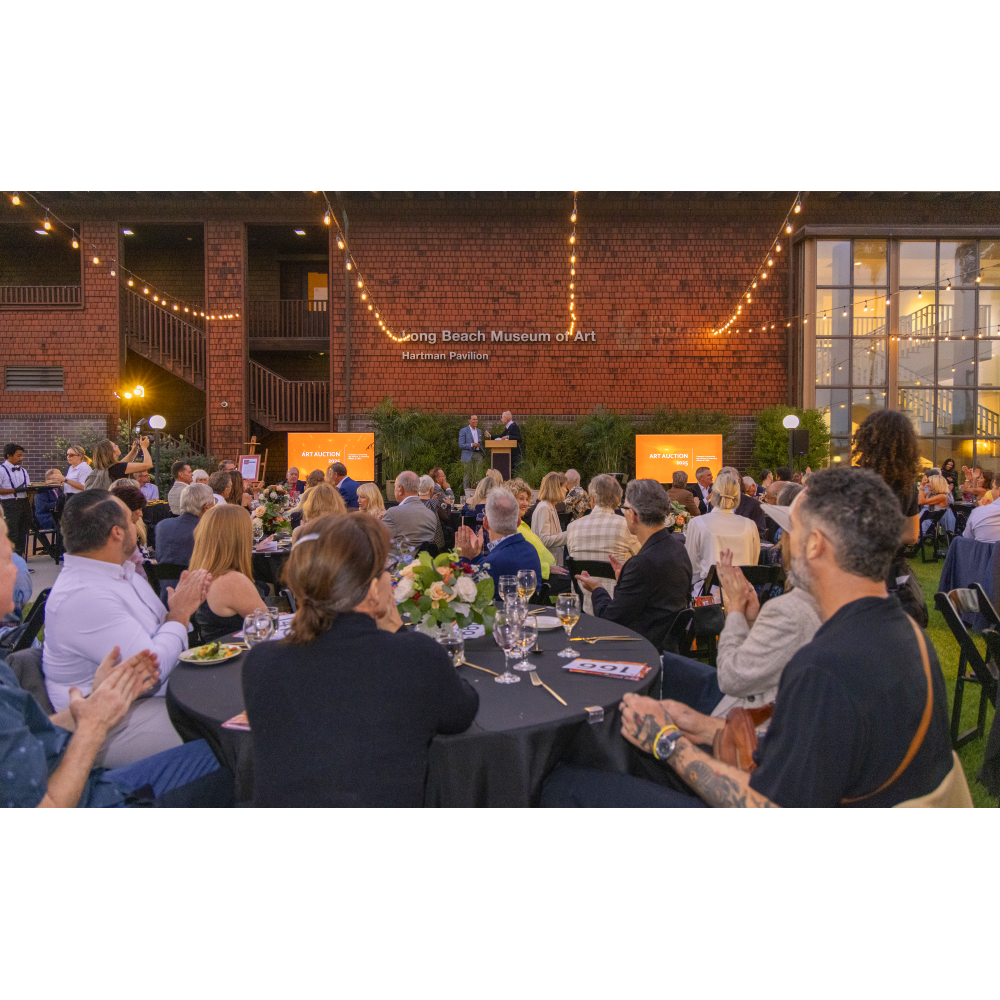Guests seated at round tables outdoors during an evening event at the Long Beach Museum of Art. String lights hang above the courtyard as attendees listen to speakers at a podium in front of the Hartman Pavilion.