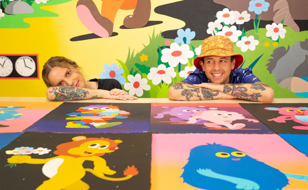 A woman and a man with tattooed arms rest their chins on their hands behind a table displaying bright, playful cartoon illustrations, with a whimsical mural of flowers and characters on the wall behind them.