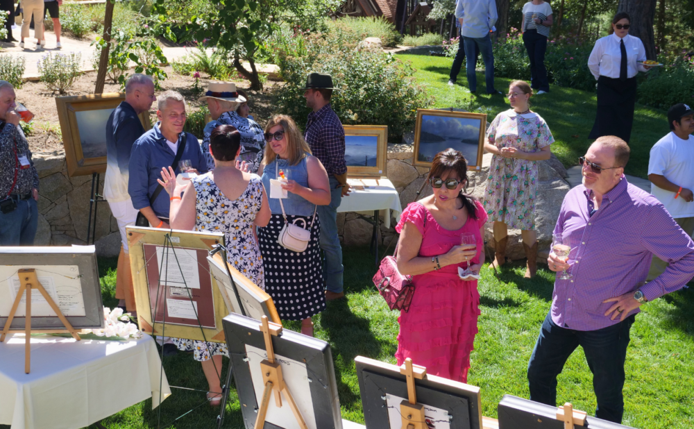 People socializing at an outdoor art display, holding drinks and conversing while viewing framed paintings arranged on easels in a garden setting.