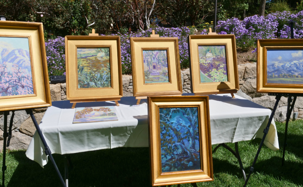 Table display of framed landscape paintings on easels, set outdoors against a stone wall with purple flowers and greenery behind.
