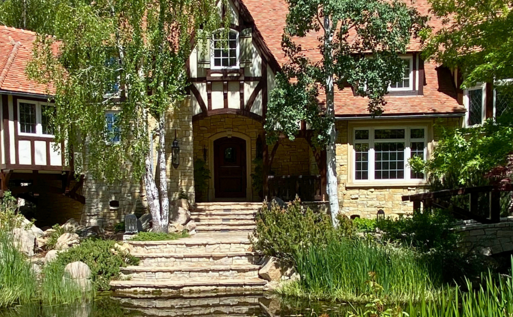 Tudor-style house with a red tile roof and timber framing, viewed from a stone stairway crossing a small pond, surrounded by lush trees and landscaped greenery.