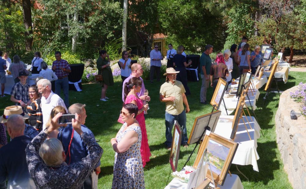 Outdoor garden event with guests mingling on a lawn, viewing framed artworks displayed on easels and tables, with servers and trees in the background.