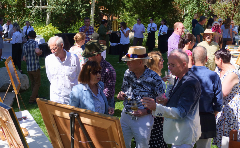 A group of people gathered outside looking at art and having a conversation. Background includes various trees and plants.