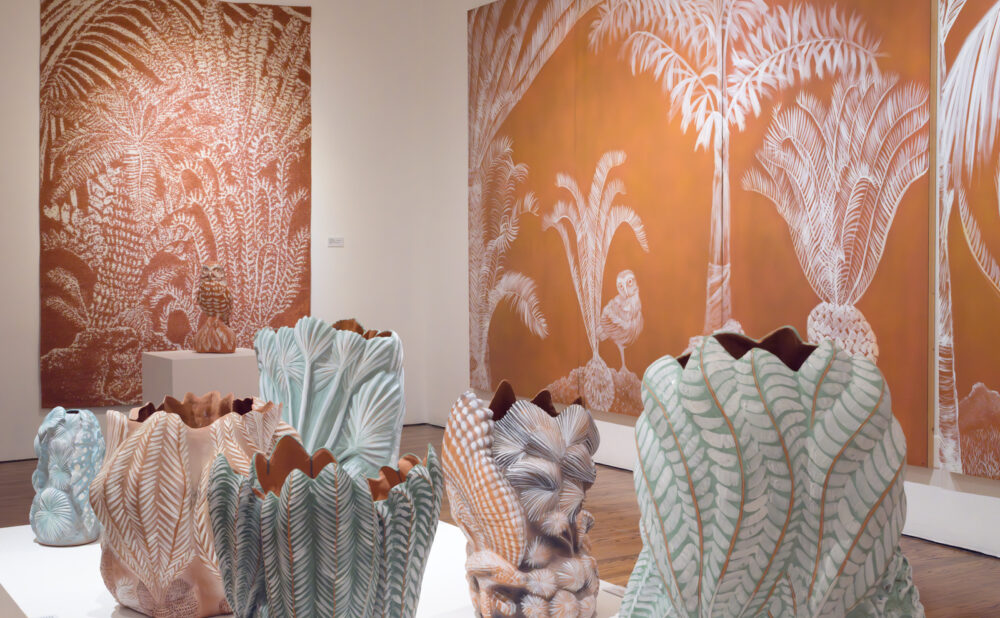 Art gallery installation featuring large orange-and-white wall panels with illustrated palm trees, birds, and foliage, alongside several sculptural ceramic vessels in soft pastel tones (green, peach, and blue) shaped like layered leaves or shells, displayed on a central platform against a wooden floor.