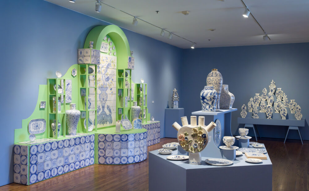Gallery exhibition featuring blue-and-white ceramic artworks displayed on pastel green and blue pedestals and shelving, including vases, plates, and sculptural pieces arranged around a decorative tiled wall installation under track lighting.