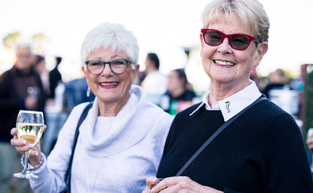 Two people wearing eyeglasses stand outdoors at a social gathering. One person in the foreground wears a dark sweater with a white collared shirt and red-framed glasses. Another person beside them wears a light-colored sweater and holds a glass of white wine. Several attendees are visible in the softly blurred background.