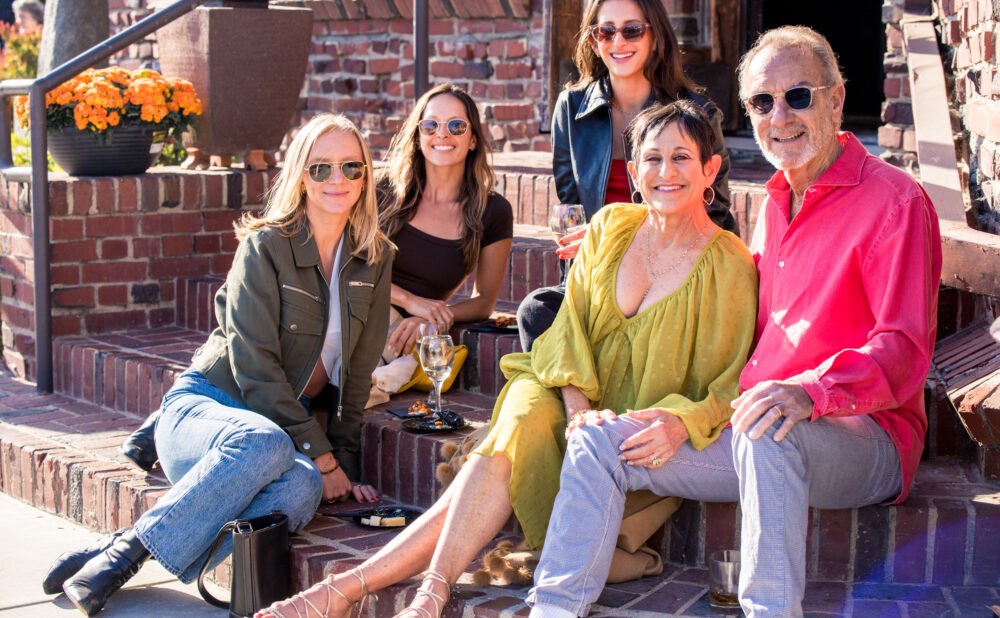 Five people wearing sunglasses sit together on brick steps outside a building. The group is gathered around drinks and small plates of food, with potted orange flowers nearby. Bright sunlight illuminates the scene, creating a casual outdoor social setting.