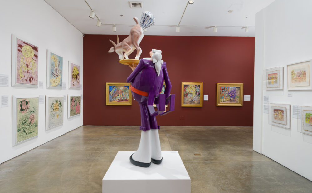 A brightly lit museum gallery with a large, whimsical sculpture centered on a white pedestal. The sculpture depicts a stylized purple-suited figure holding a tray above its head with a deer balancing on top. Colorful framed paintings hang on the surrounding walls, including a deep red accent wall behind the sculpture.
