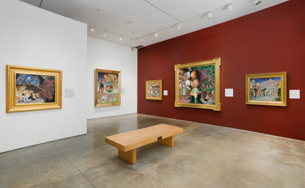 A spacious museum gallery with polished concrete floors and white walls, featuring several large, vividly detailed paintings in gold frames hung on a deep red wall. A simple wooden bench sits in the center of the room for viewing.