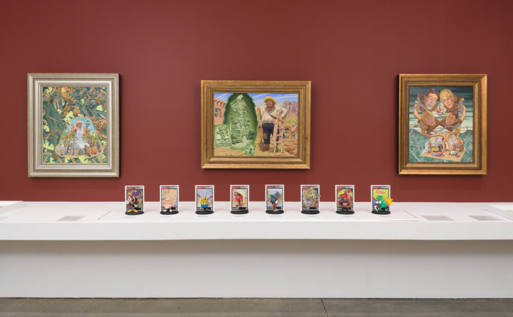 A museum display with three framed paintings mounted on a red wall above a long white display case. Inside the case are small figurines and collectible-style objects arranged in a row beneath the artworks.