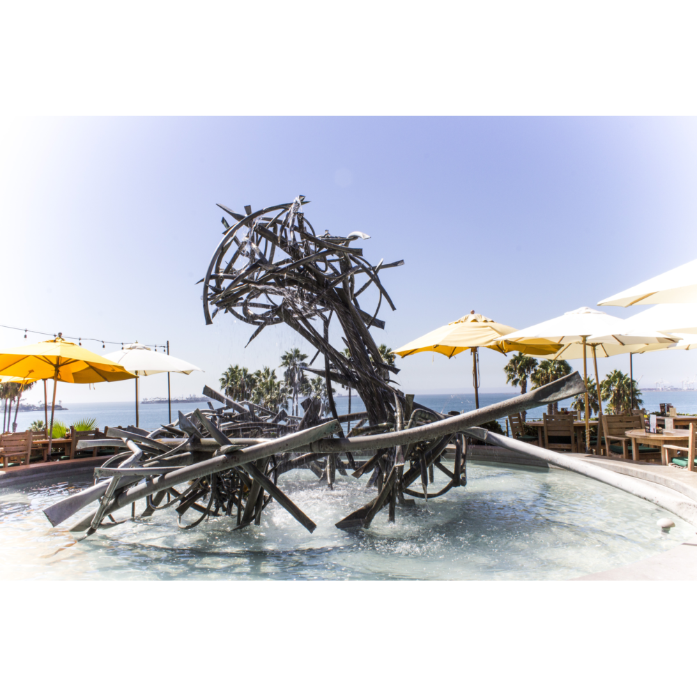 Abstract metal sculpture fountain made of tangled, curved steel rods, spraying water in multiple directions, set on a seaside patio with white and yellow umbrellas, wooden tables and chairs, palm trees, and a clear blue sky over the ocean in the background.