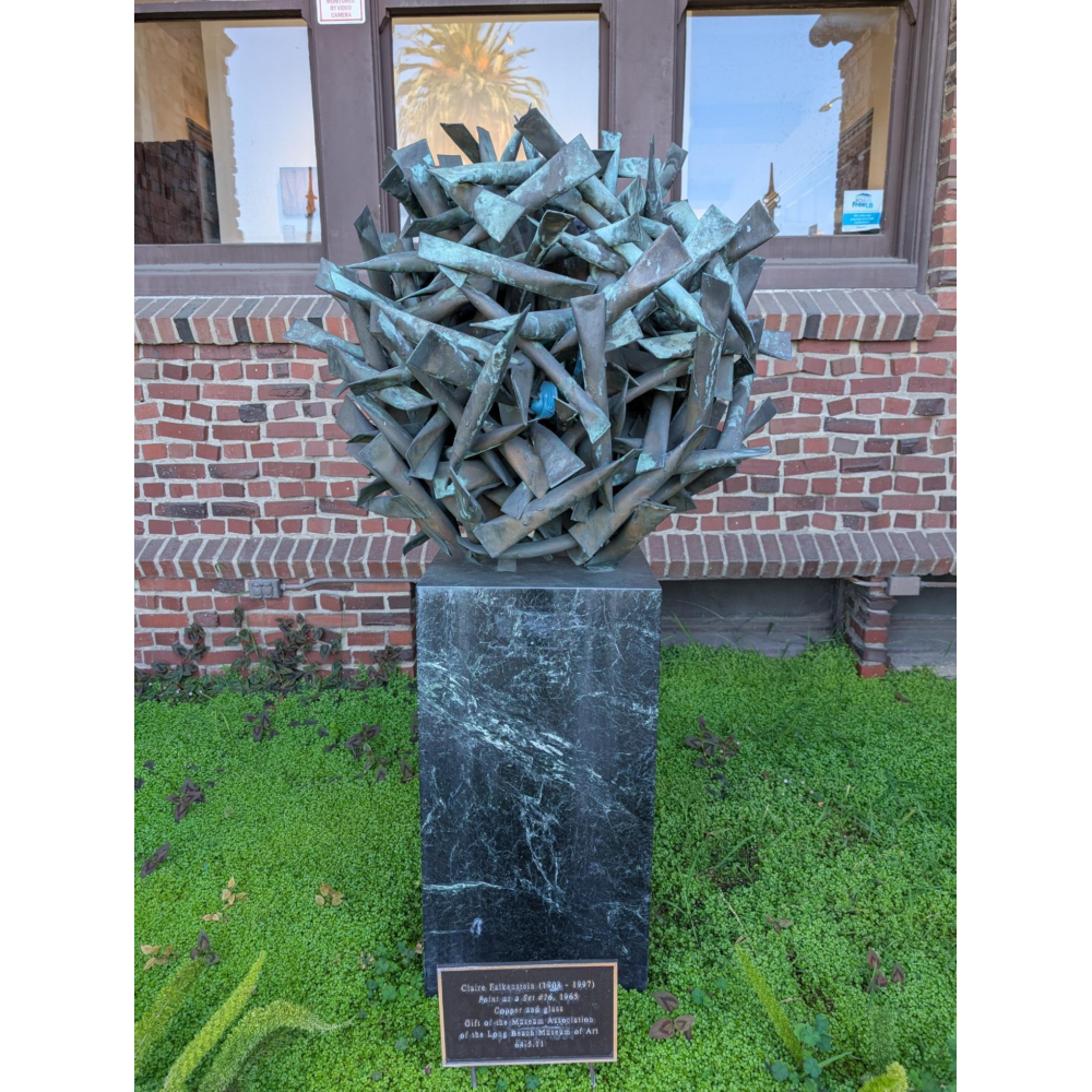 Round abstract sculpture made of clustered, spiky metal tubes with a green patina, mounted on a dark stone pedestal, set against a brick wall with large windows reflecting a palm tree and sky.
