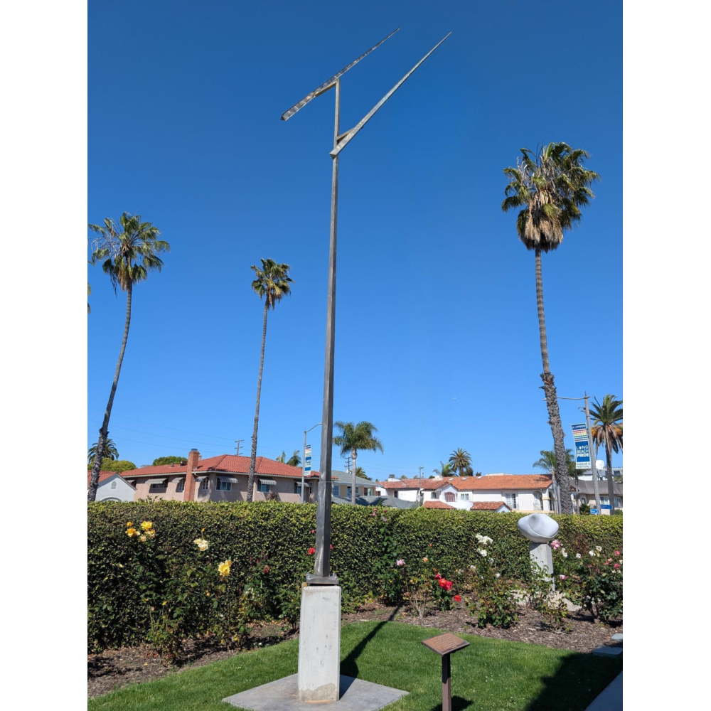 Tall, minimalist metal sculpture with two long, angled blades at the top mounted on a slender pole, standing on a concrete base in a landscaped garden with blooming rose bushes, palm trees, and low houses under a clear blue sky.