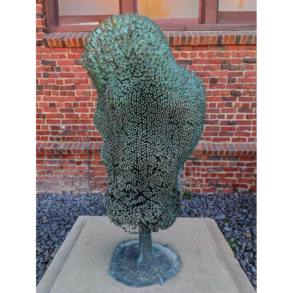 Abstract green-patina metal sculpture made of densely packed, short rod-like elements forming an organic, coral- or plant-like shape, mounted on a pedestal in front of a red brick wall with windows and a bed of dark stones at the base.