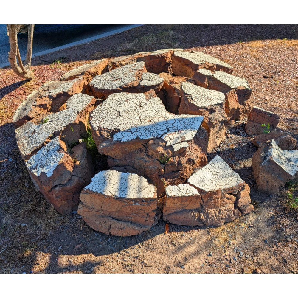 Circular, low sculpture made of rough, reddish-brown clay or stone segments arranged like a broken ring, with cracked, pale surfaces on top resembling dried earth, set on the ground in a landscaped area beside a road.