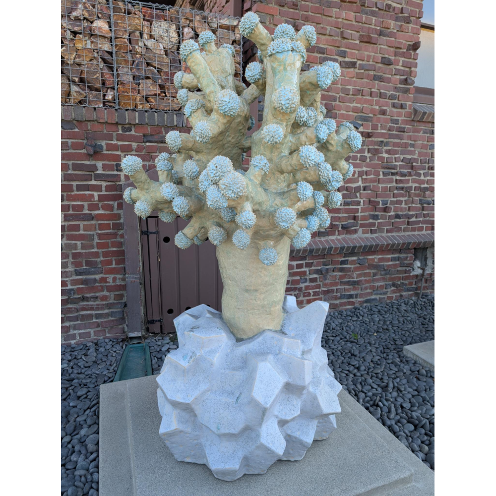 Abstract, tree-like sculpture with thick branching forms topped by many small, rounded, spiky clusters in a pale green glaze, rising from a faceted white base, set on a stone pedestal in front of a red brick wall with a metal rock-filled cage feature to the side.
