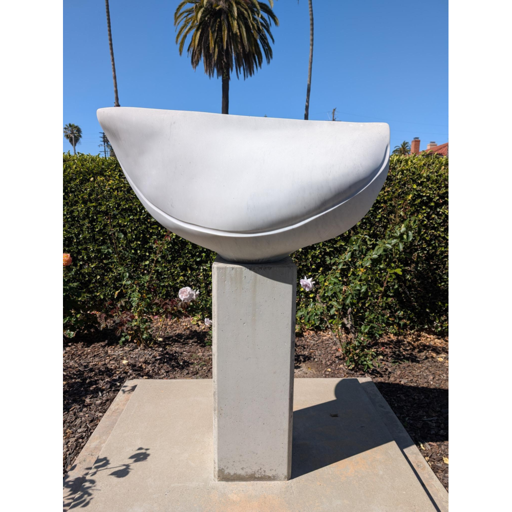 A smooth white abstract sculpture shaped like a shallow curved bowl sits on a rectangular concrete pedestal in a sunny garden, with a hedge, pink flowers, and tall palm trees in the background.