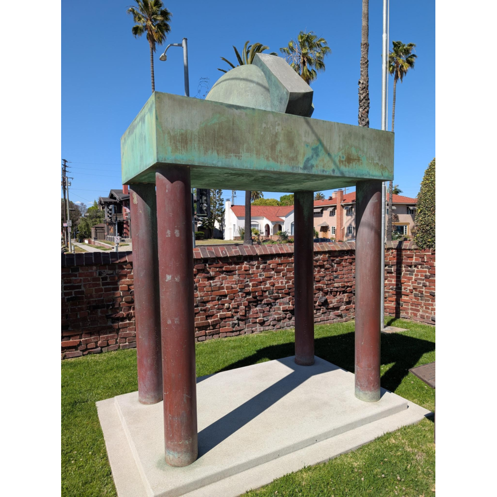 Large abstract metal sculpture with a weathered green patina, consisting of a rectangular slab supported by four cylindrical columns, topped with geometric forms, installed on a concrete base in a grassy area beside a brick wall, with palm trees, houses, and a clear blue sky in the background.