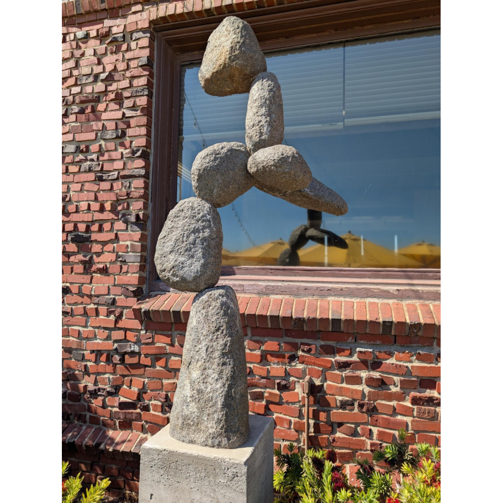 Abstract stacked stone sculpture composed of several rounded, irregular granite forms balanced vertically on a pedestal, set against a red brick wall with a window reflecting yellow umbrellas and outdoor seating.