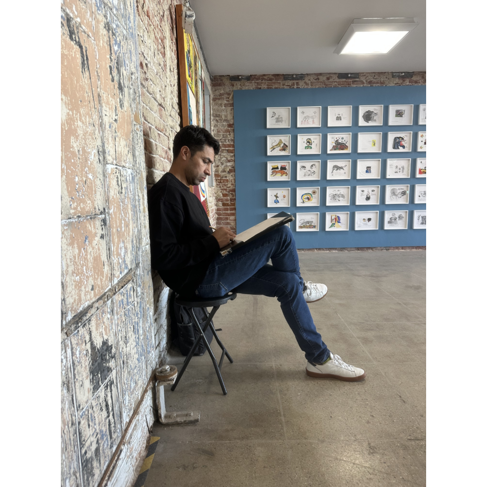 A person sketches while seated on a folding chair during Downtown Draws at LBMA Downtown, surrounded by exposed brick walls and gallery displays featuring framed drawings and artwork.