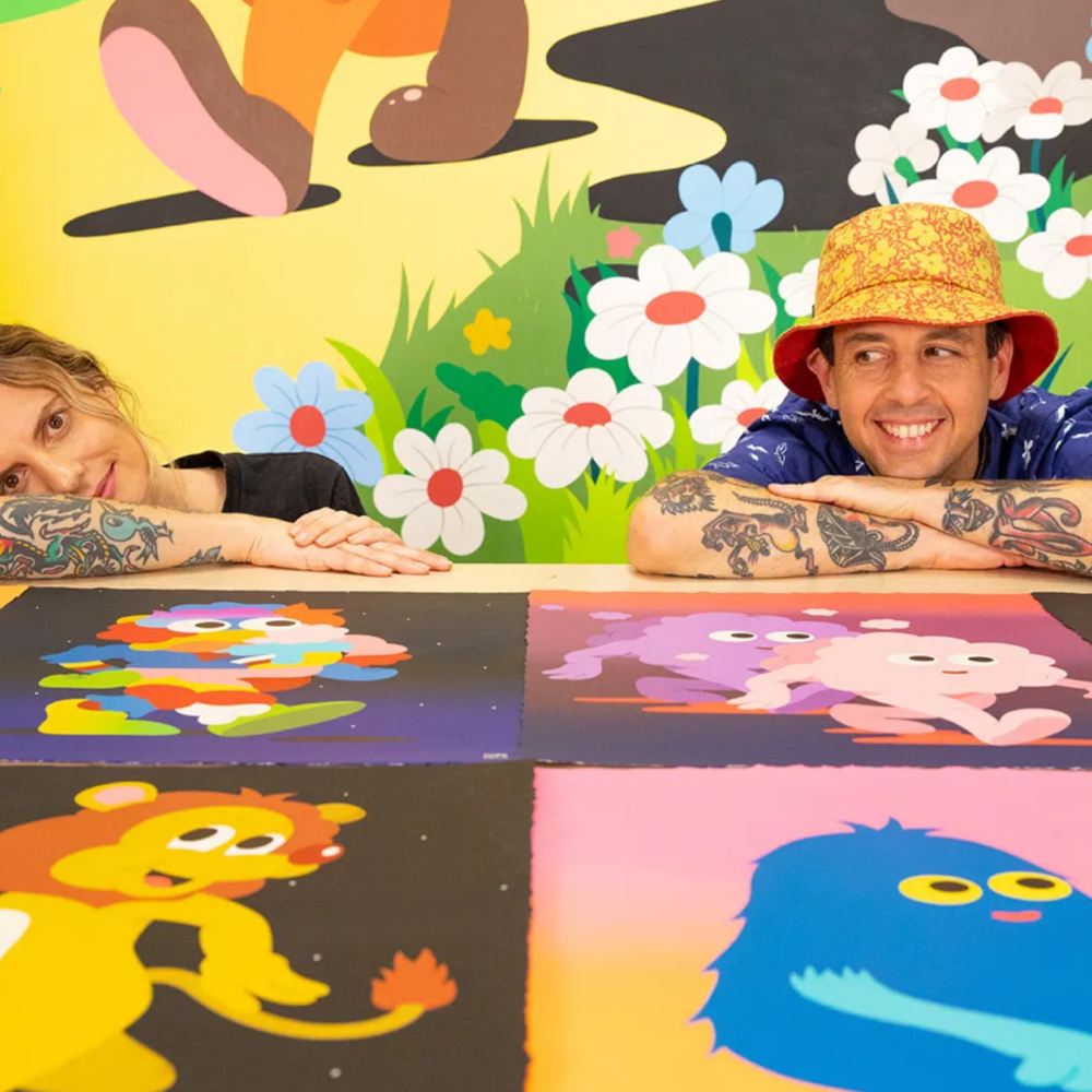 A woman and a man with tattooed arms rest their chins on their hands behind a table displaying bright, playful cartoon illustrations, with a whimsical mural of flowers and characters on the wall behind them.